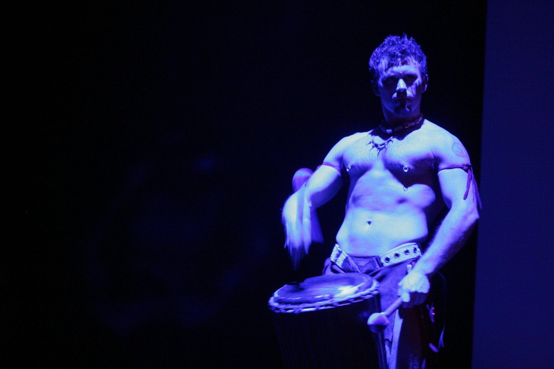 A shirtless man is playing a drum in a dark room.