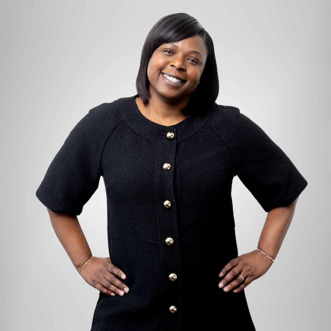 Smiling woman in a black buttoned dress posing with hands on hips against a gray background