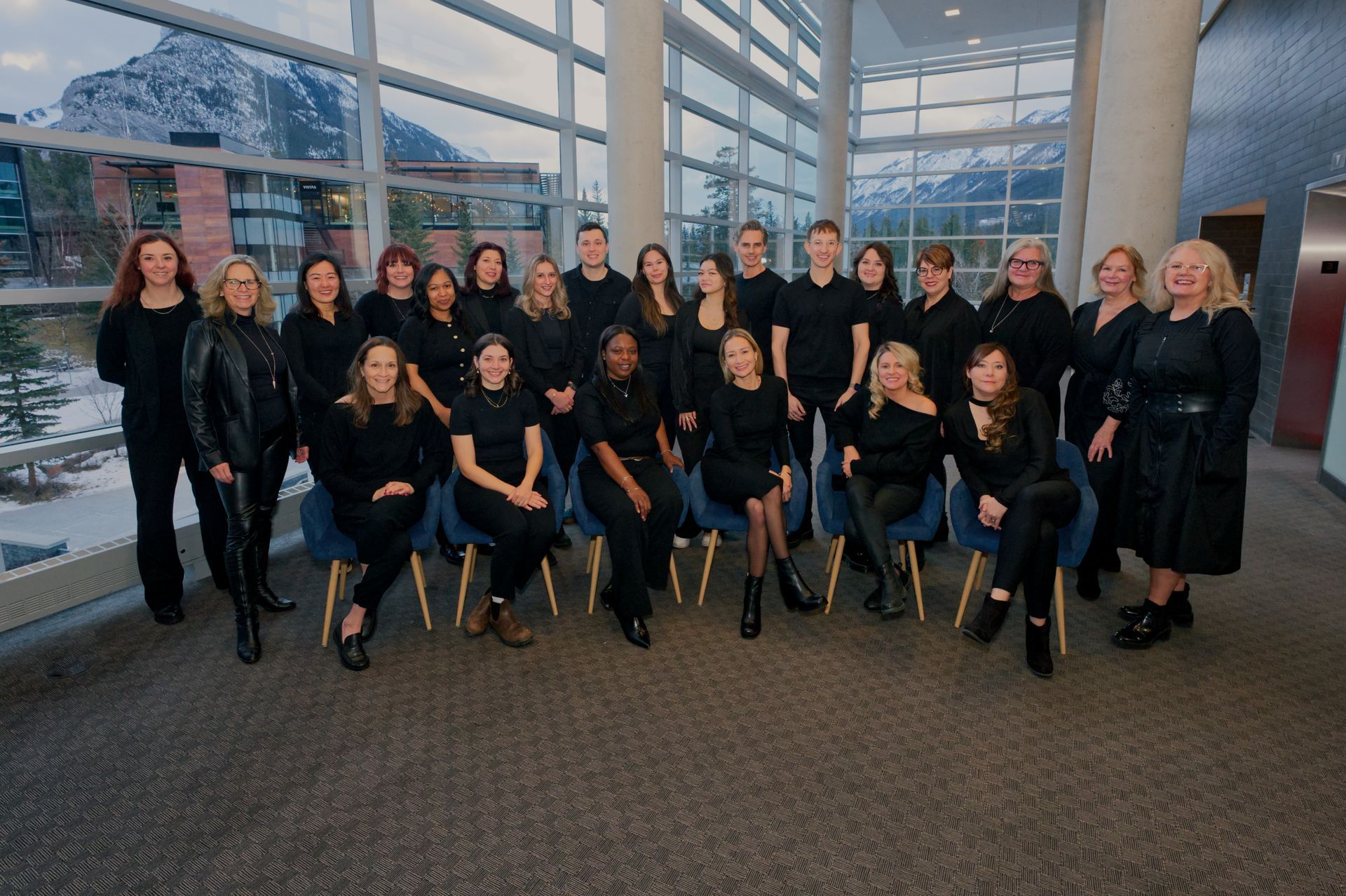 Group of people in black outfits posing indoors, with large windows overlooking mountains.