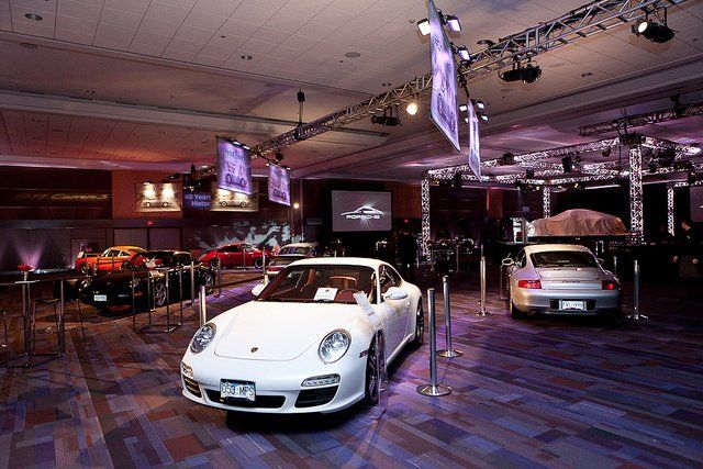 White Porsche sports car on display at an auto show, surrounded by other vehicles and spotlights.