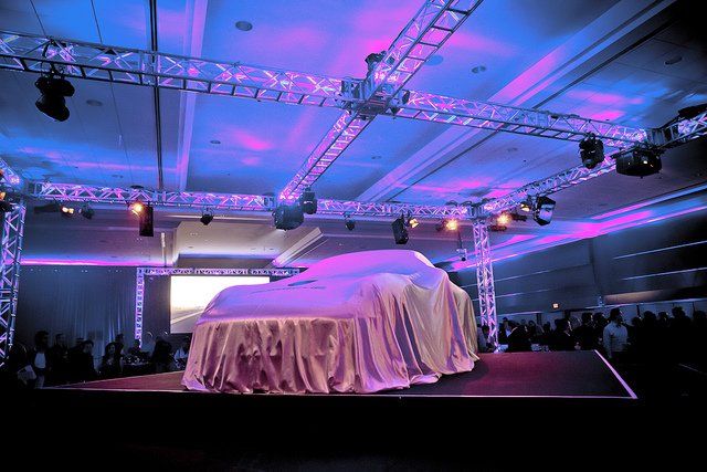 Car covered by a sheet on a stage at an event, with stage lights and an audience.