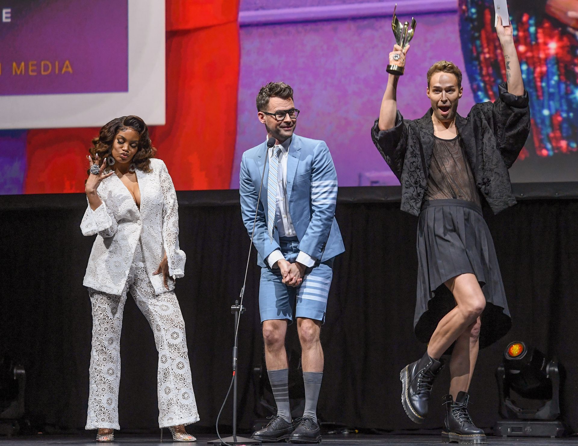 Three presenters on stage, one holding up a trophy, another clapping, with colorful event lighting behind them