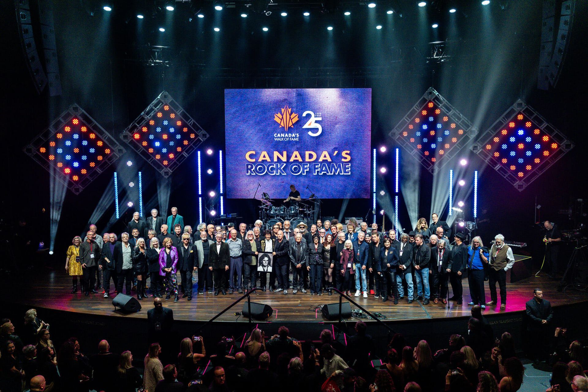 A large group of people are standing on a stage in front of a large screen that says canada 's rock of fame.