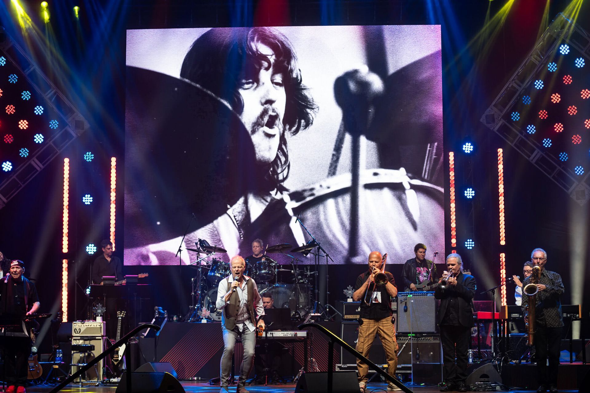 A group of people on a stage with a large screen behind them showing a man playing drums