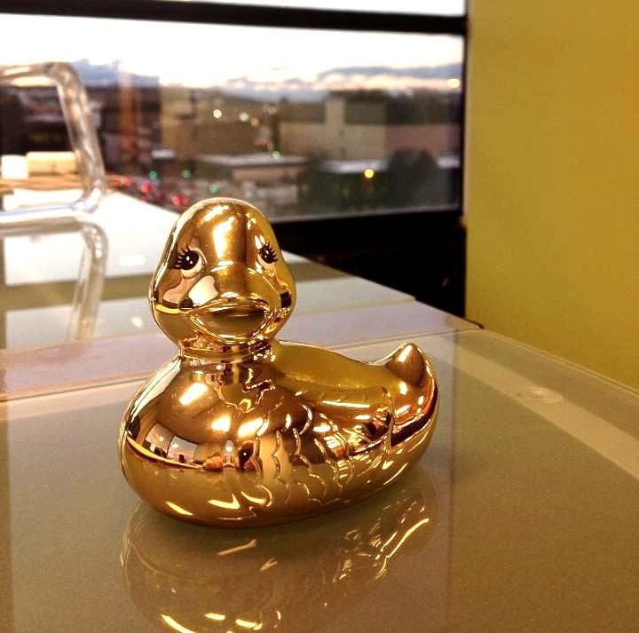 A gold rubber duck sits on a table in front of a window