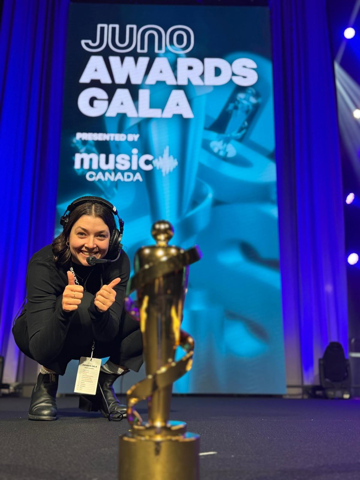 Woman kneeling next to Juno award trophy, smiling with thumbs up at the Juno Awards Gala backdrop.