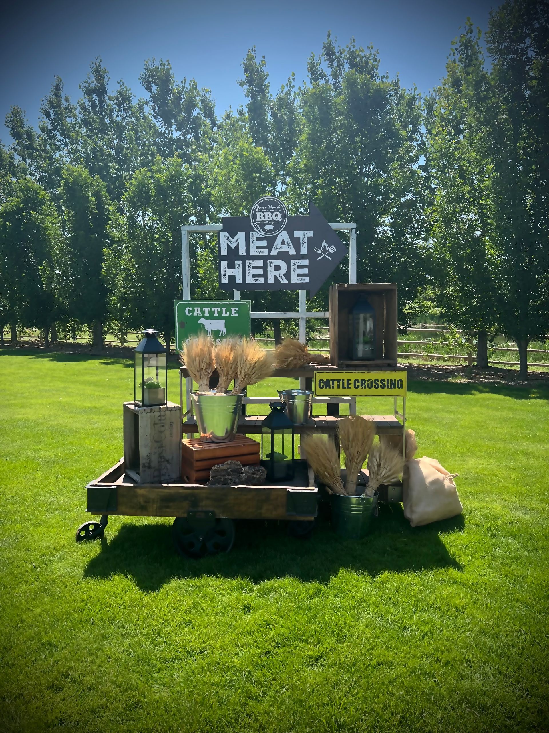 Meat & Here sign on a rustic display cart with hay, metal buckets, and wooden crates, set on green grass with trees.