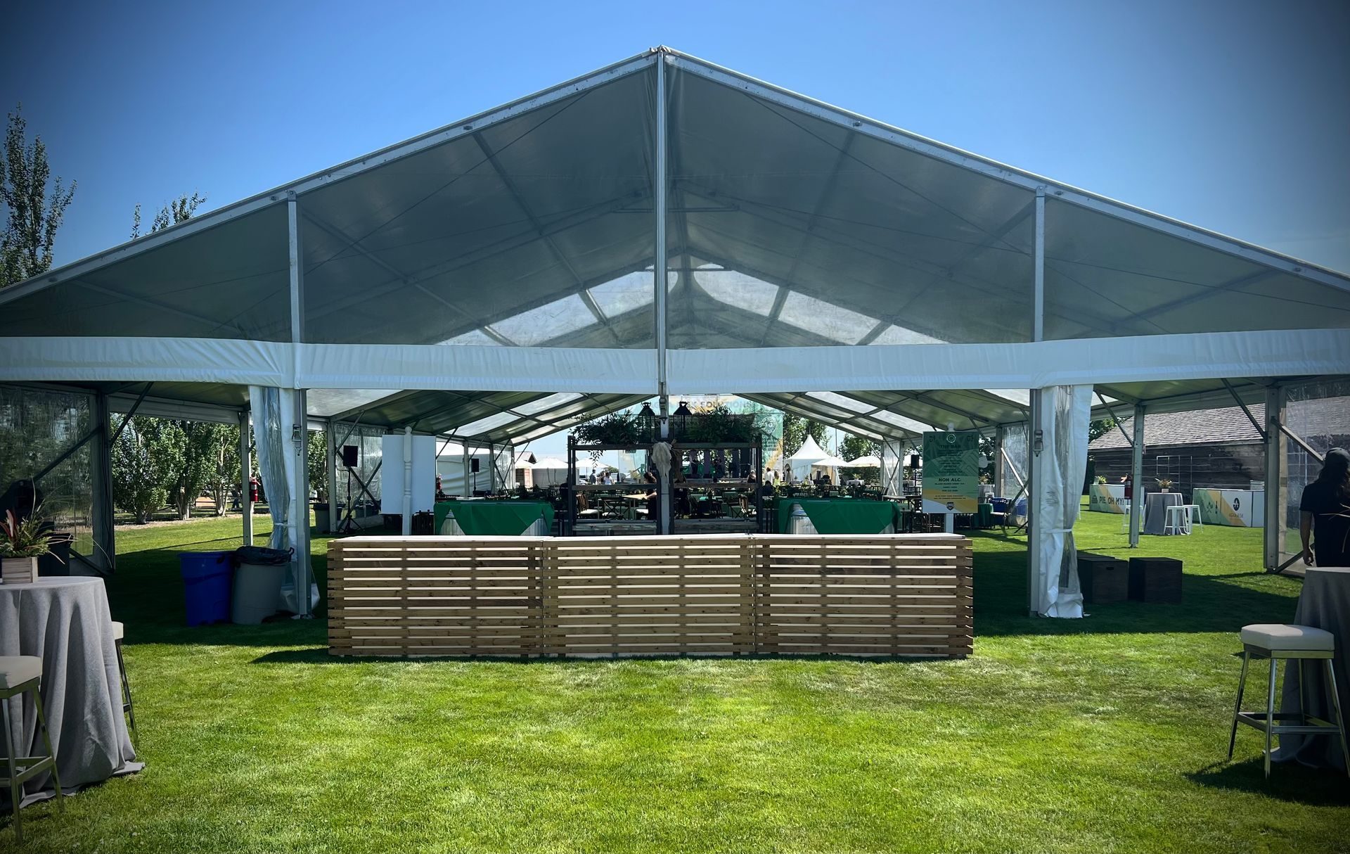 Clear tent set up on grass with wooden bar and tables, sunny day.