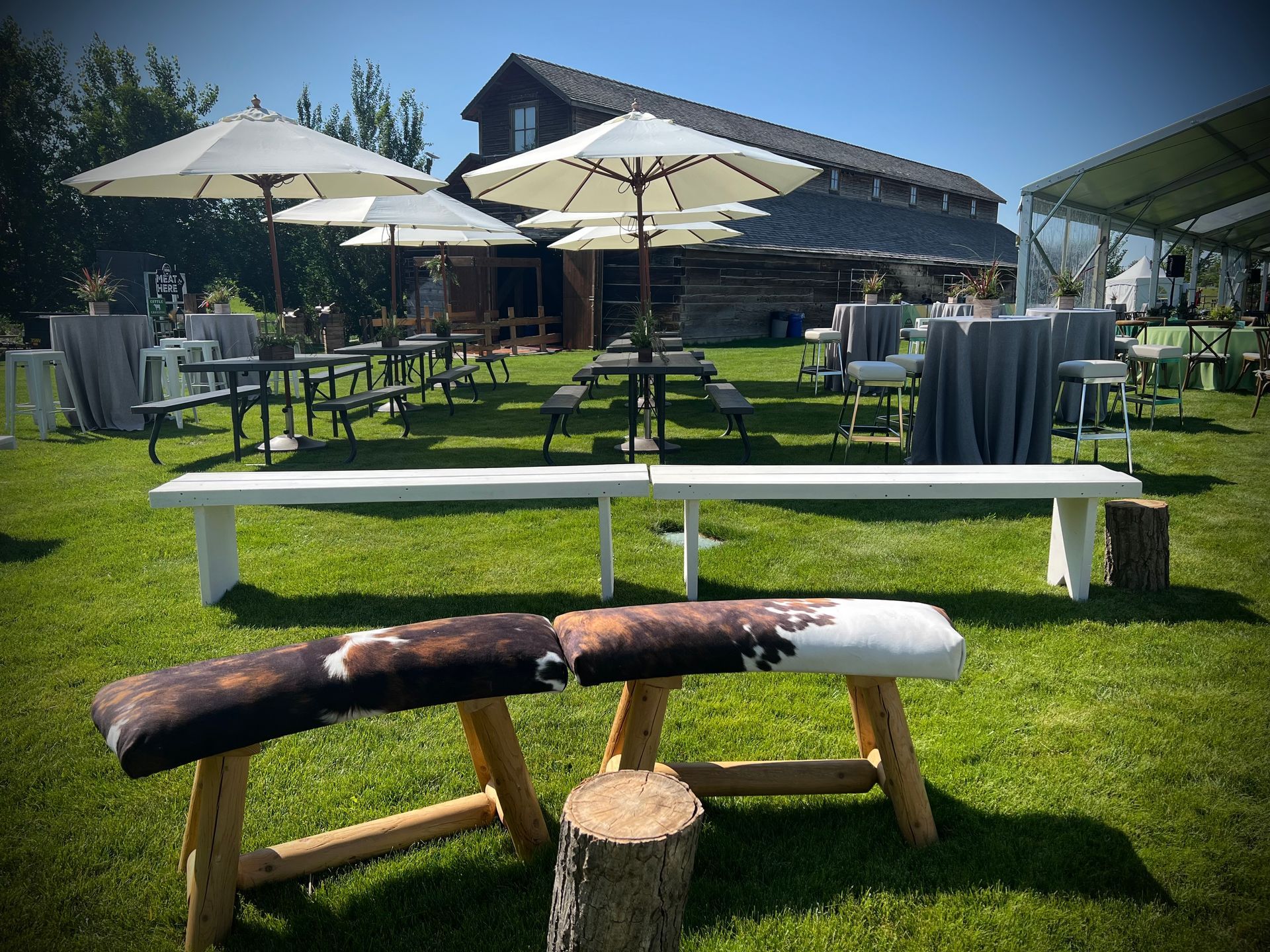 Outdoor event setting: benches, tables, umbrellas, a barn, and a tent on a grassy lawn.