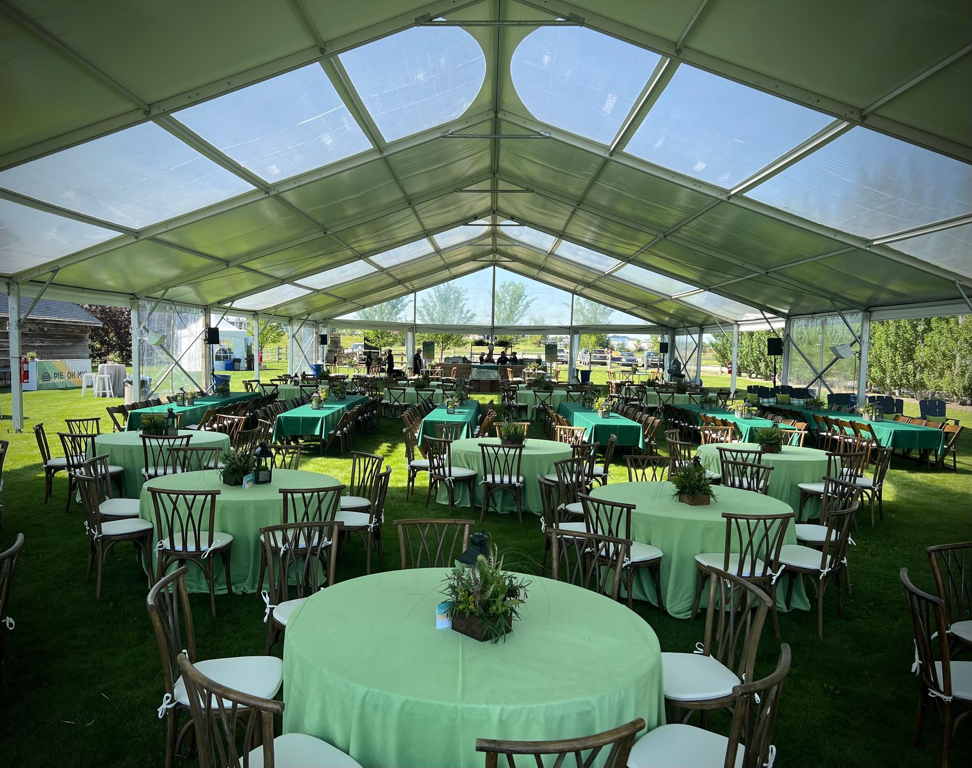 A clear-roofed tent set up on grass, filled with round tables covered in green linens and wooden chairs, for an event.