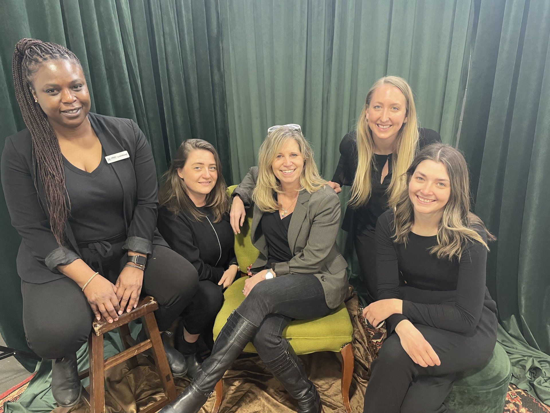 A group of women are sitting on chairs in front of a green curtain.