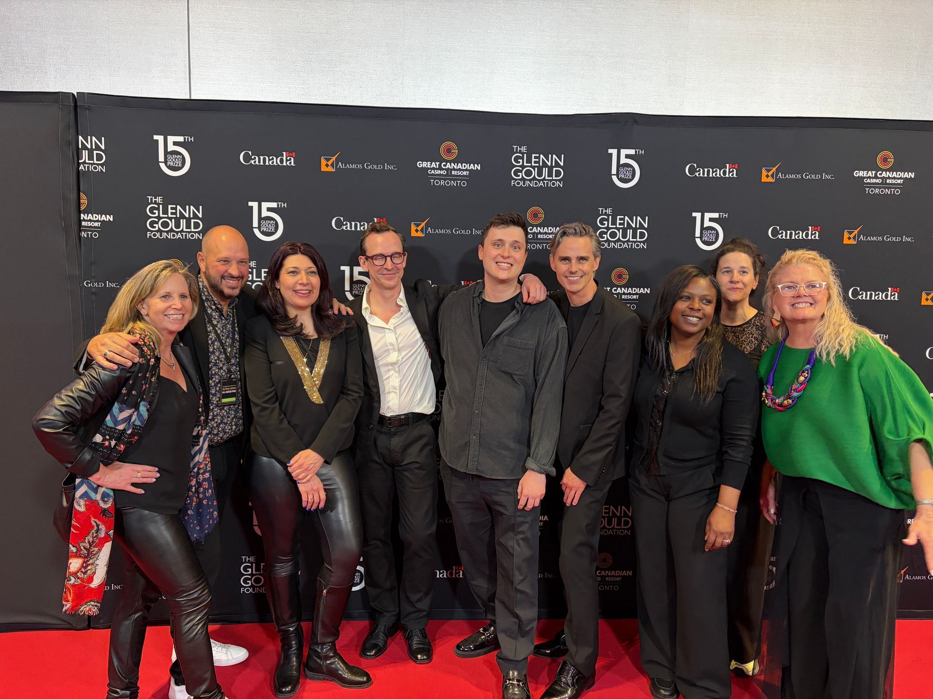 Group of eight people posing on a red carpet in front of a step-and-repeat backdrop, smiling and dressed in black and green.