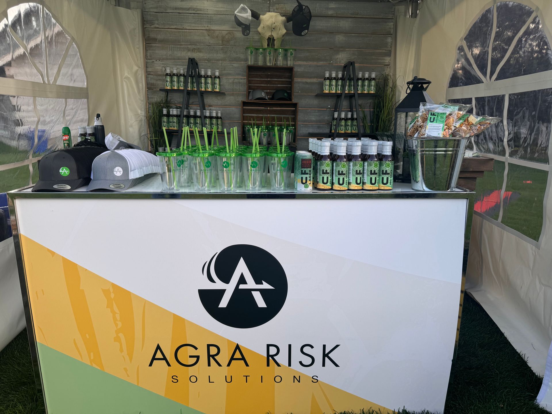 Bar setup for Agra Risk Solutions with products, hats, and glasses in a tent.