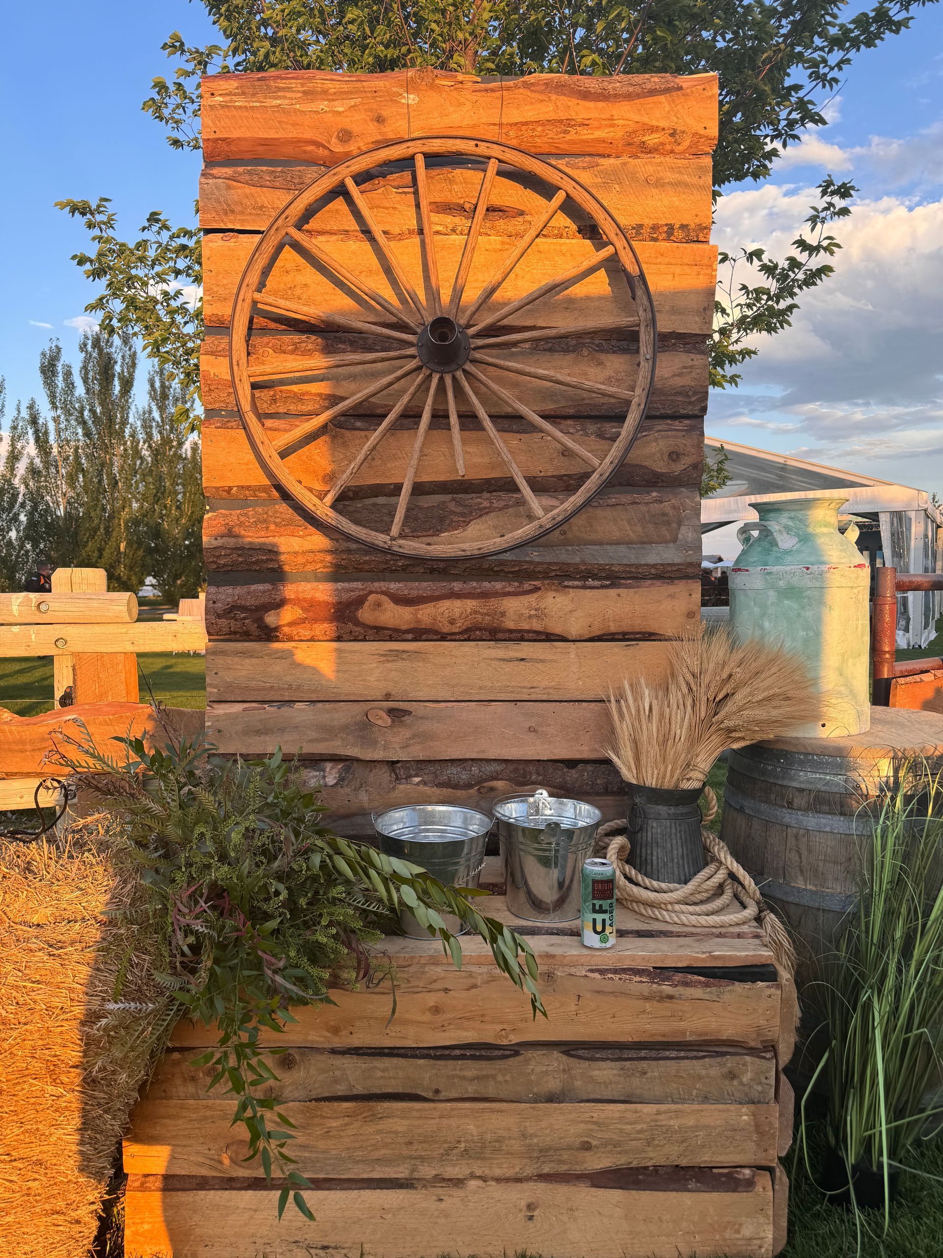 Rustic wooden display with a wagon wheel, metal buckets, and wheat. Outdoor setting.