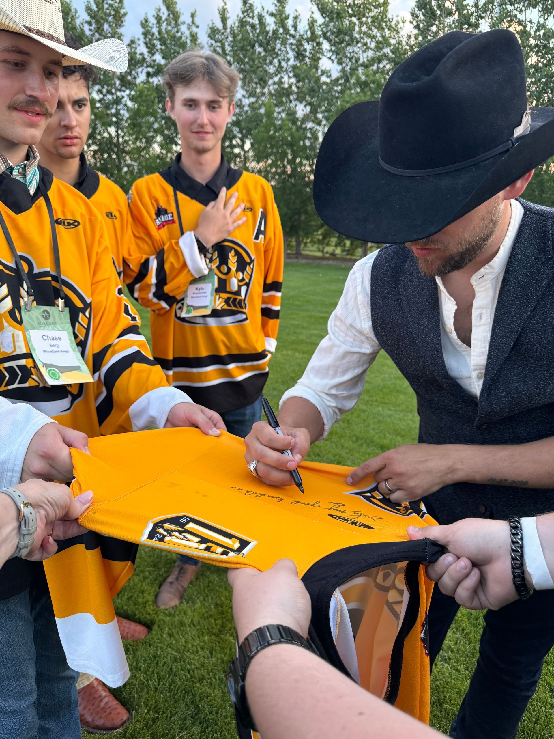 Man in cowboy hat signing hockey jersey held by young men in yellow jerseys outdoors.