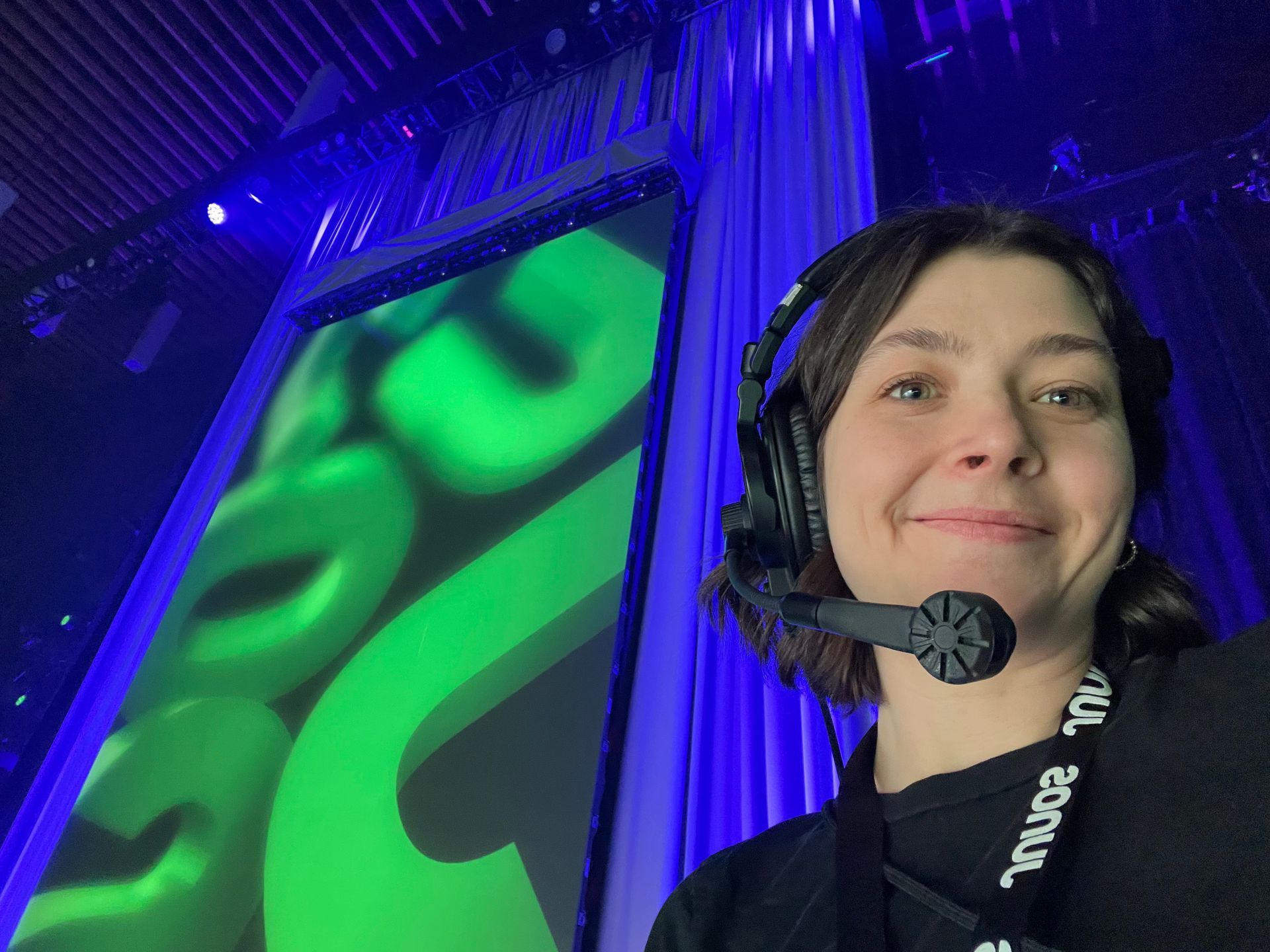 Woman with headset smiles in front of a green graphic screen, in a venue with blue lighting.