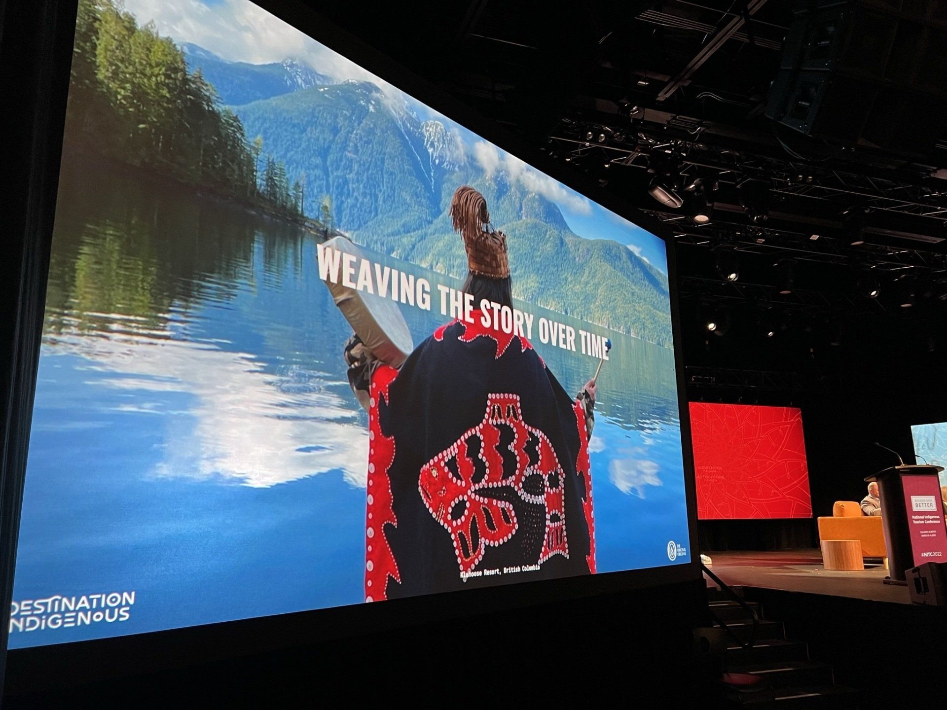 A large screen shows a man weaving a blanket in front of a lake