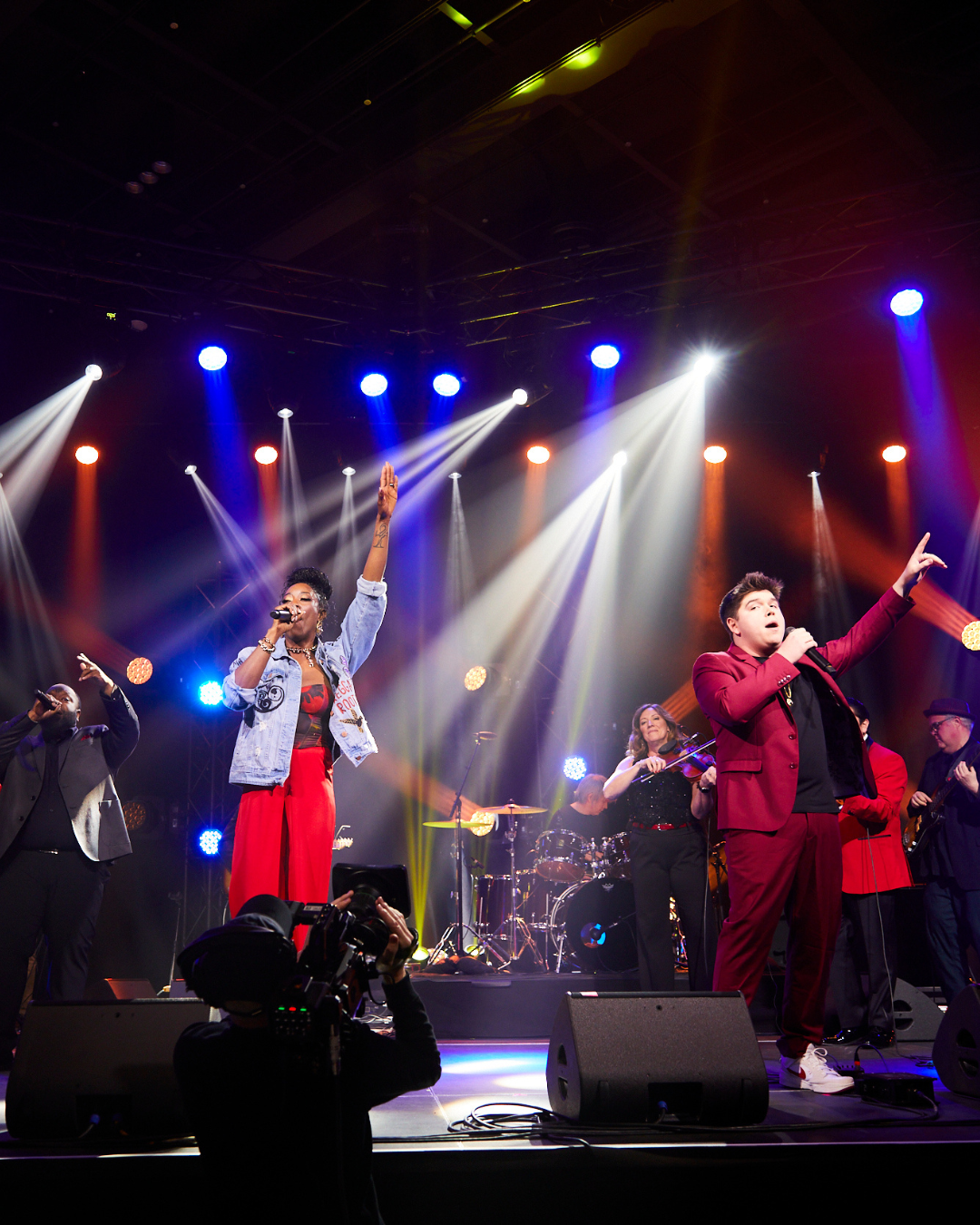 Singers performing onstage under colorful concert lights, with microphone stands and drums in the background