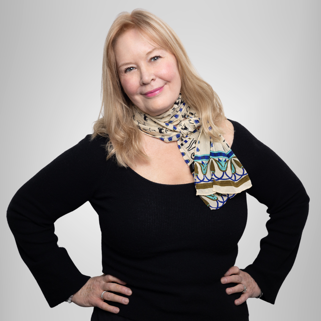 Smiling woman in a black top and patterned scarf, standing with hands on hips against a light background