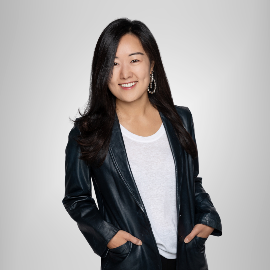 Smiling woman in a dark jacket and white shirt, standing against a light gray background with hands in pockets