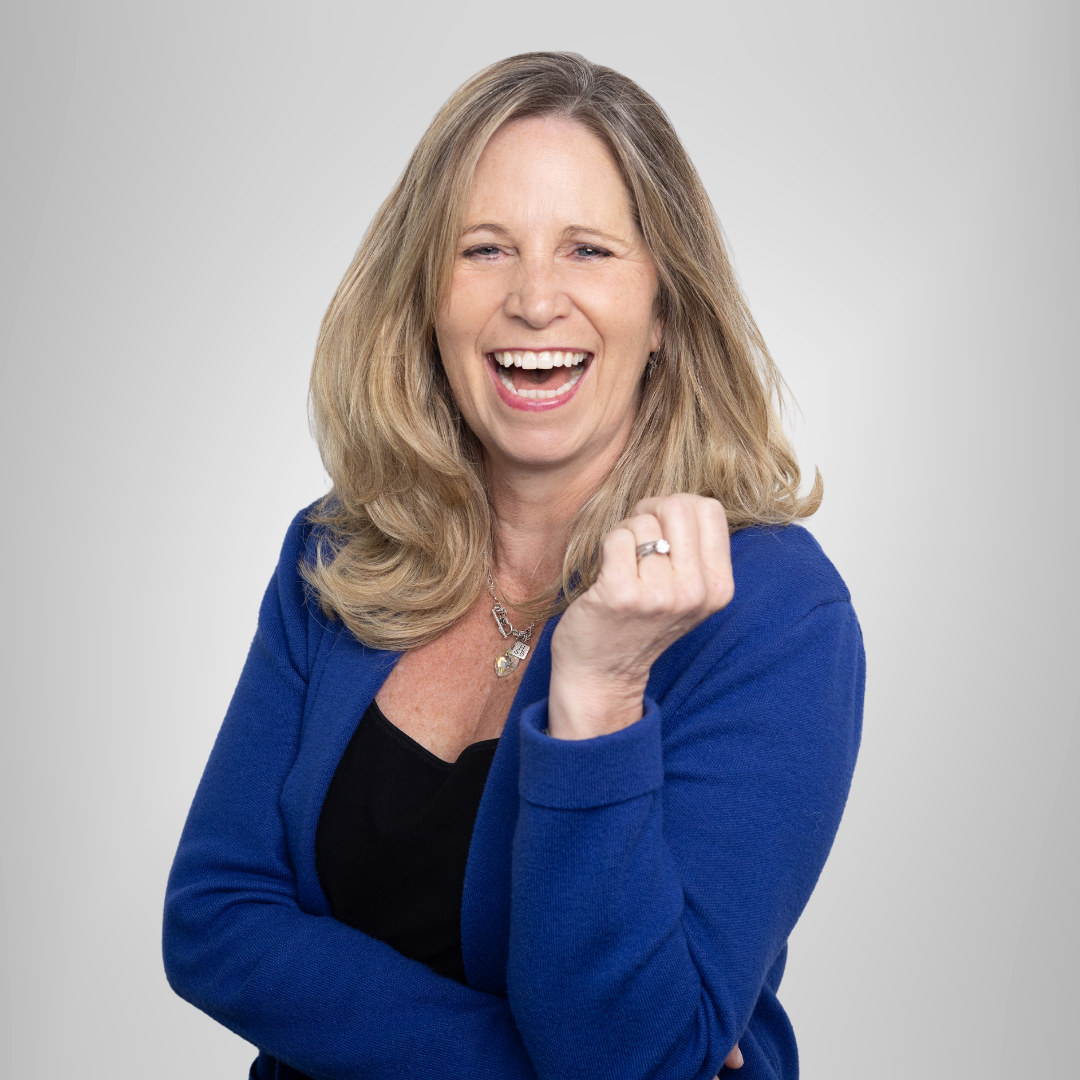 Smiling woman in a blue blazer, standing with a raised fist against a light gray background.