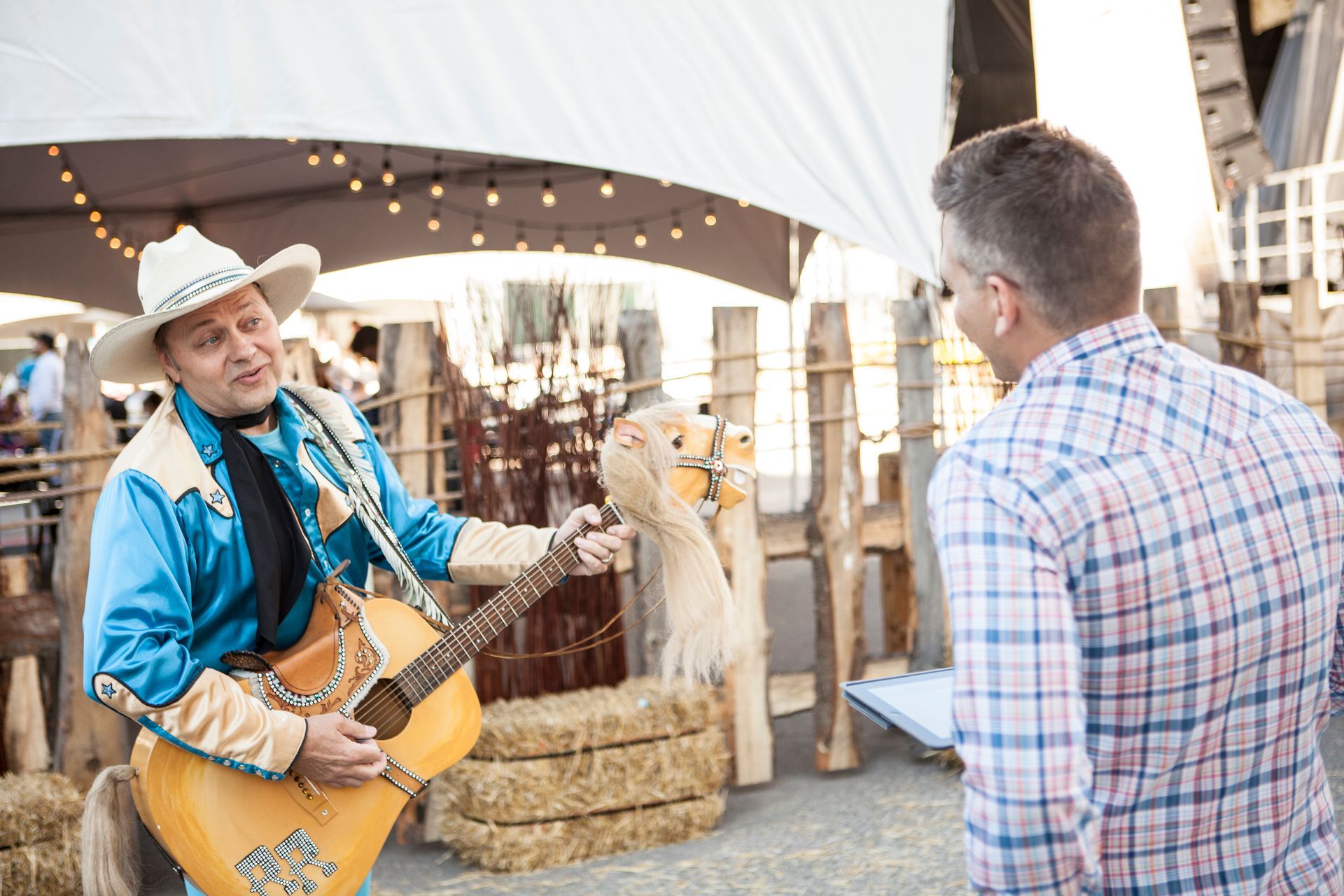 A man in a cowboy outfit is playing a guitar and talking to another man.