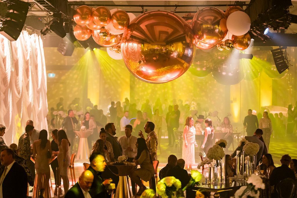 Party scene with gold balloons, people mingling, and yellow and white lighting.
