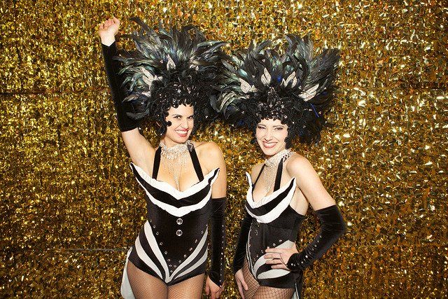 Two women in cabaret costumes are standing next to each other in front of a gold background.