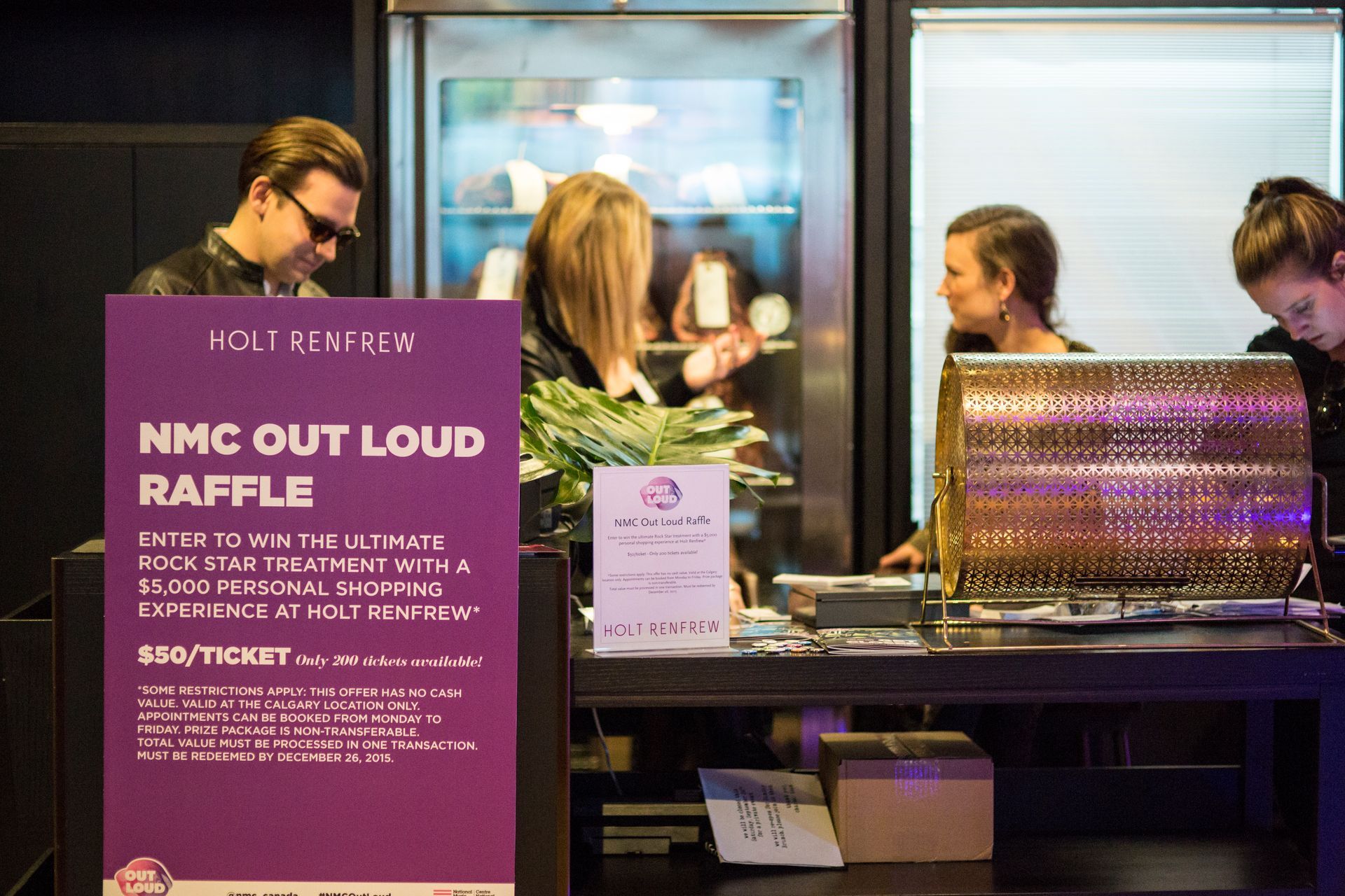 A group of people are standing around a table in front of a sign that says nmc out loud raffle.