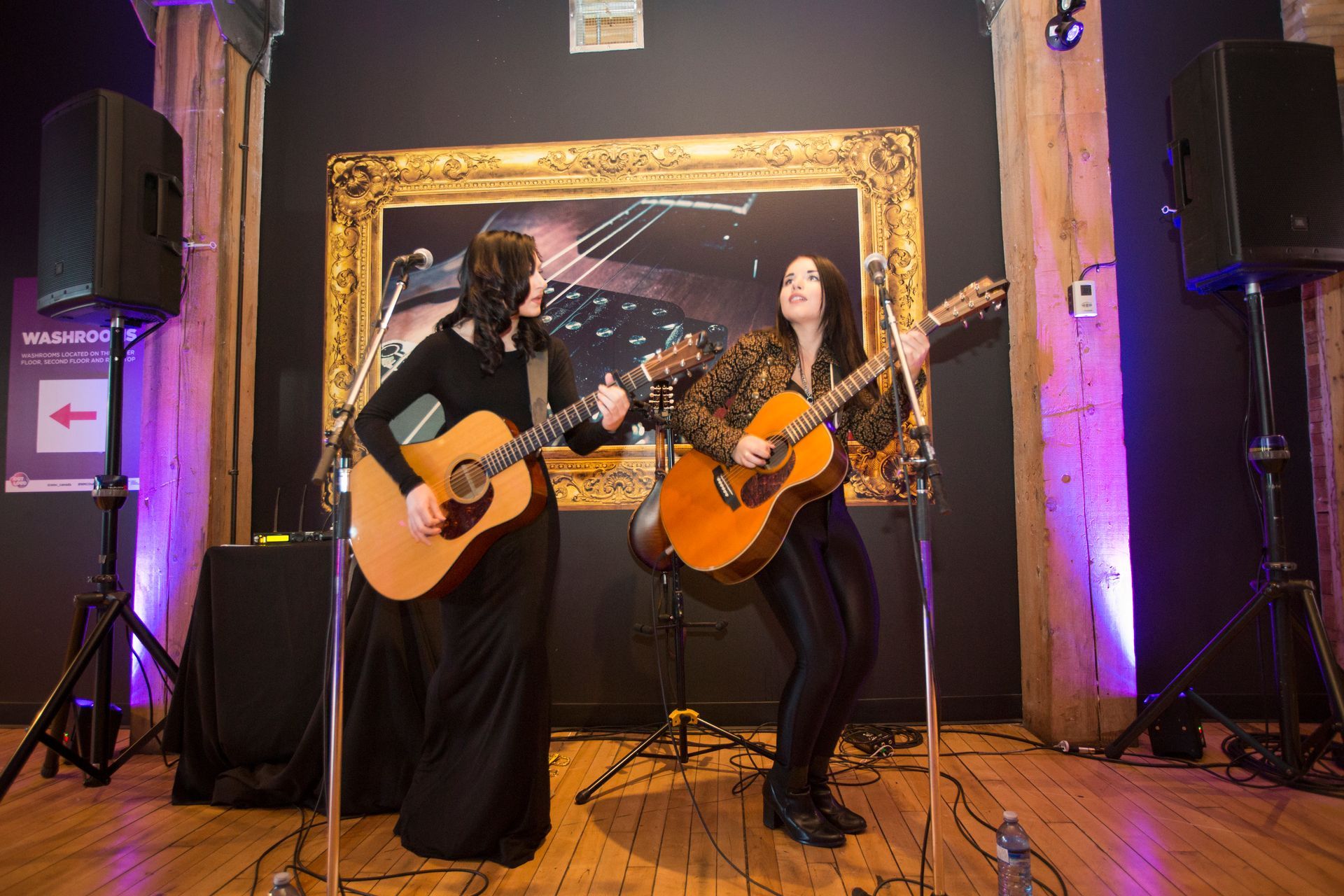 Two women are playing guitars on a stage in front of a painting.