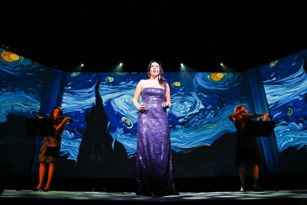 A woman in a purple dress is singing on a stage while two women play violins.