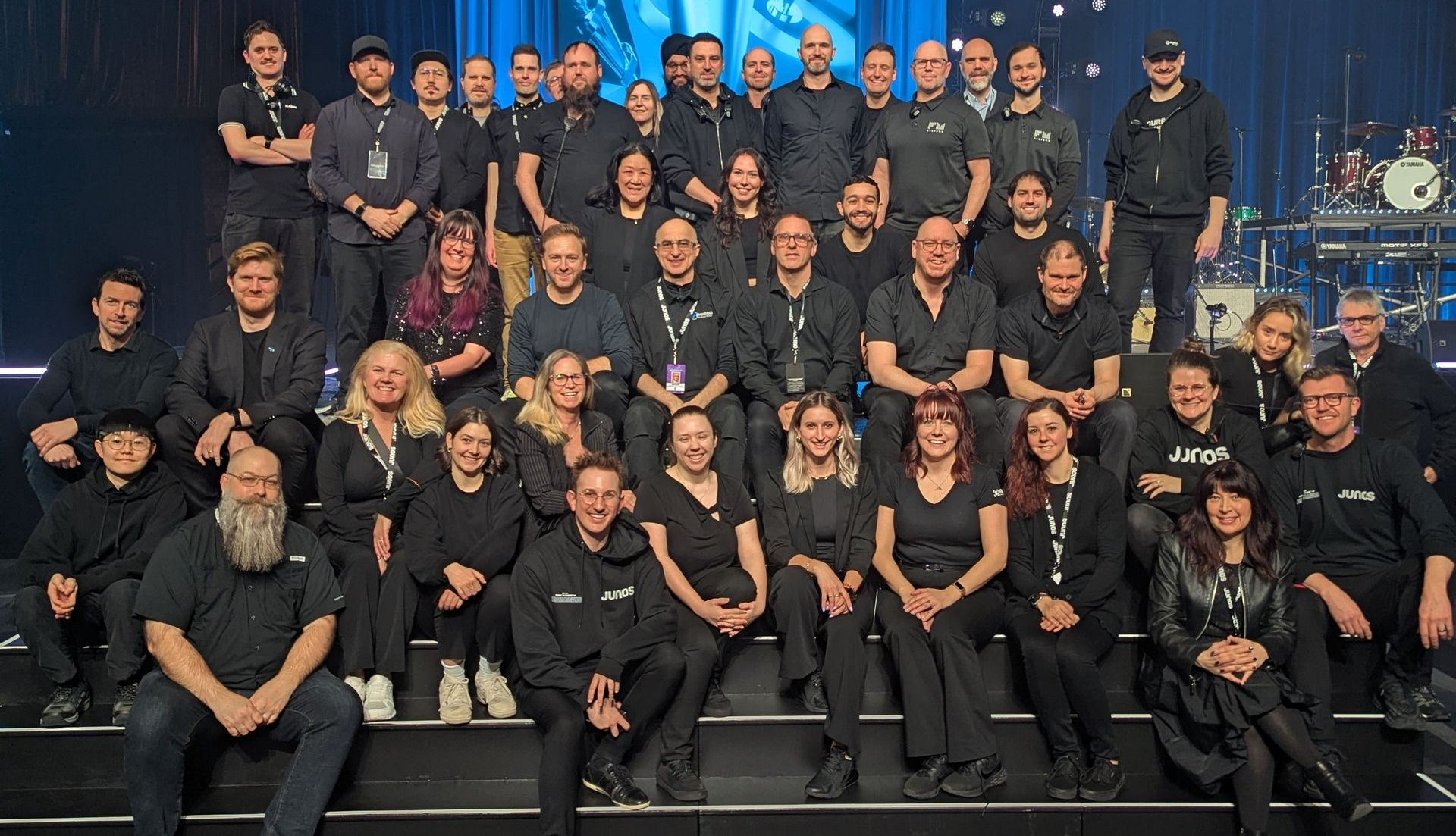 Group of people in black clothing posing on stage with musical equipment in the background.