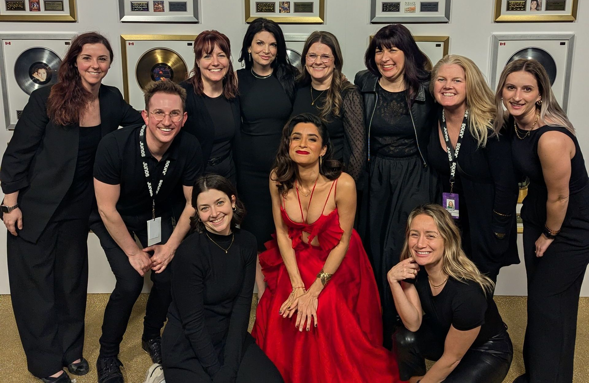 Group posing with a woman in a red dress. They stand/kneel near gold records on a wall.