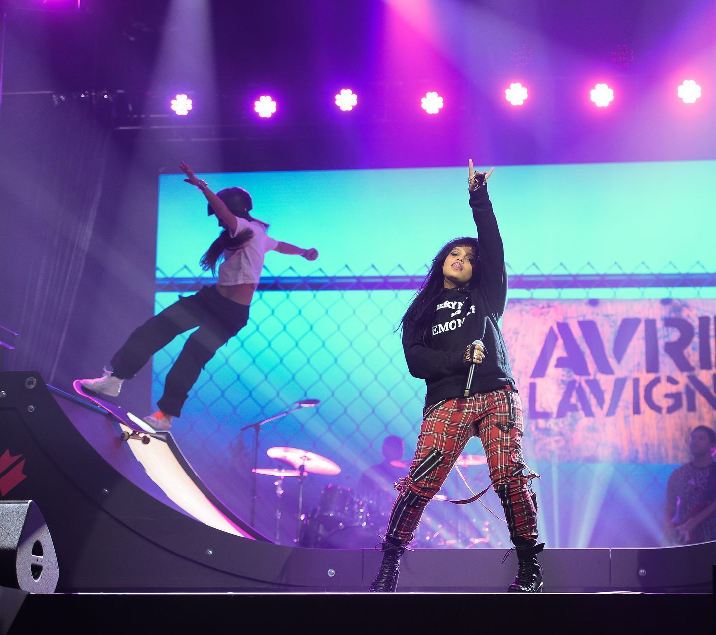 Concert performance with two dancers onstage under purple lights, one on a skateboard mid-jump