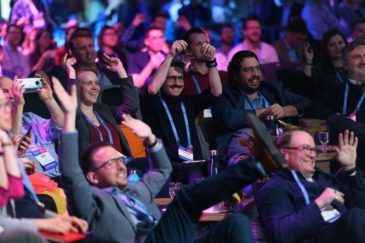 Audience cheering at an event; hands raised, smiling faces, indoor setting with blue and pink lighting.
