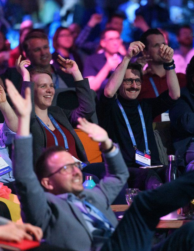 Audience members at a conference smiling and raising hands under colorful stage lighting