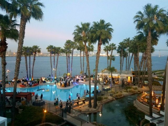Poolside event with palm trees, water, and bridge under a dusky sky, with people socializing.