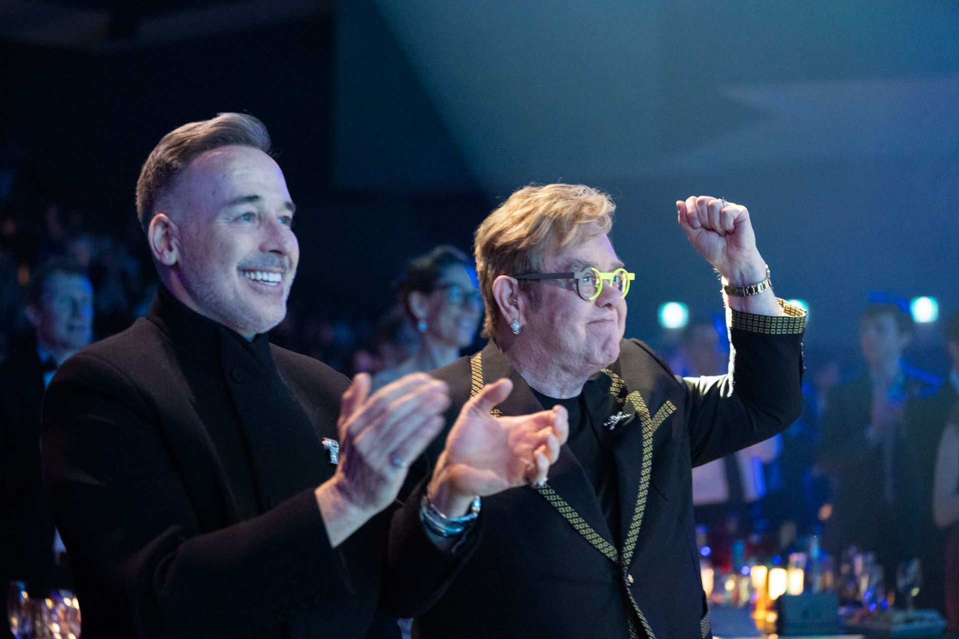 Two men smiling and clapping at a dimly lit event, with one raising a fist in celebration