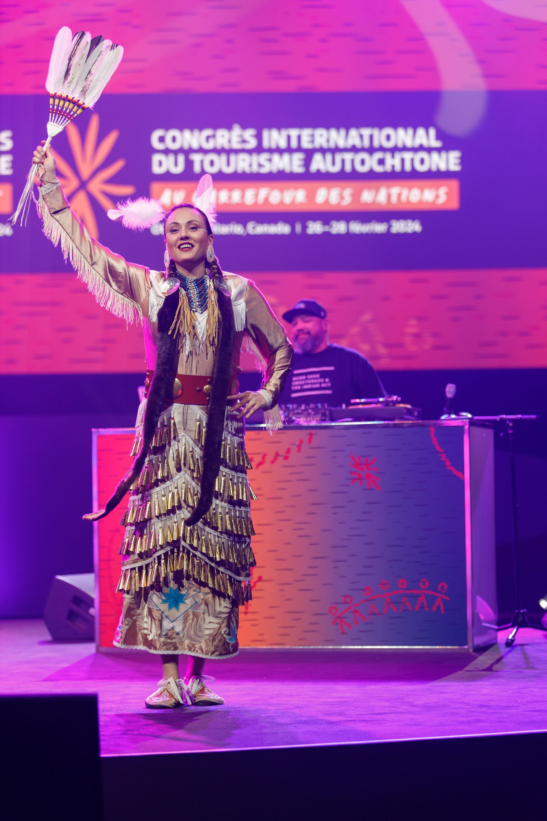 Traditional dancer performing on stage at a tourism congress, with a purple event backdrop and DJ booth behind.