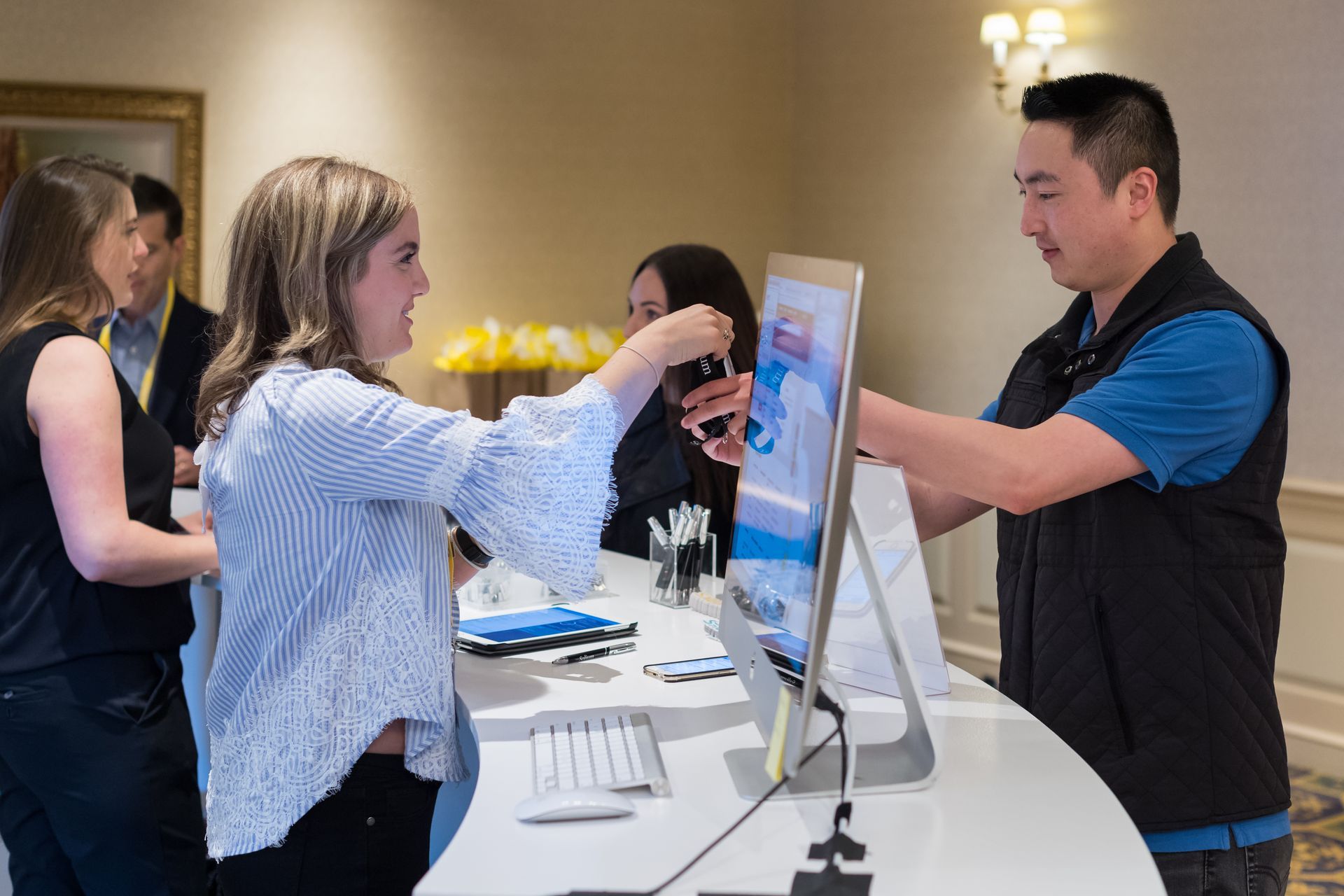 People at a registration desk: woman handing a keycard to a man, a computer, and other people in the background.