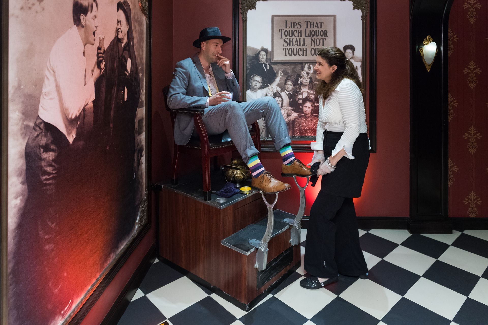 Man sits on a shoe shine stand, being attended to by a woman in a vintage-style room with black and white checkered floors.