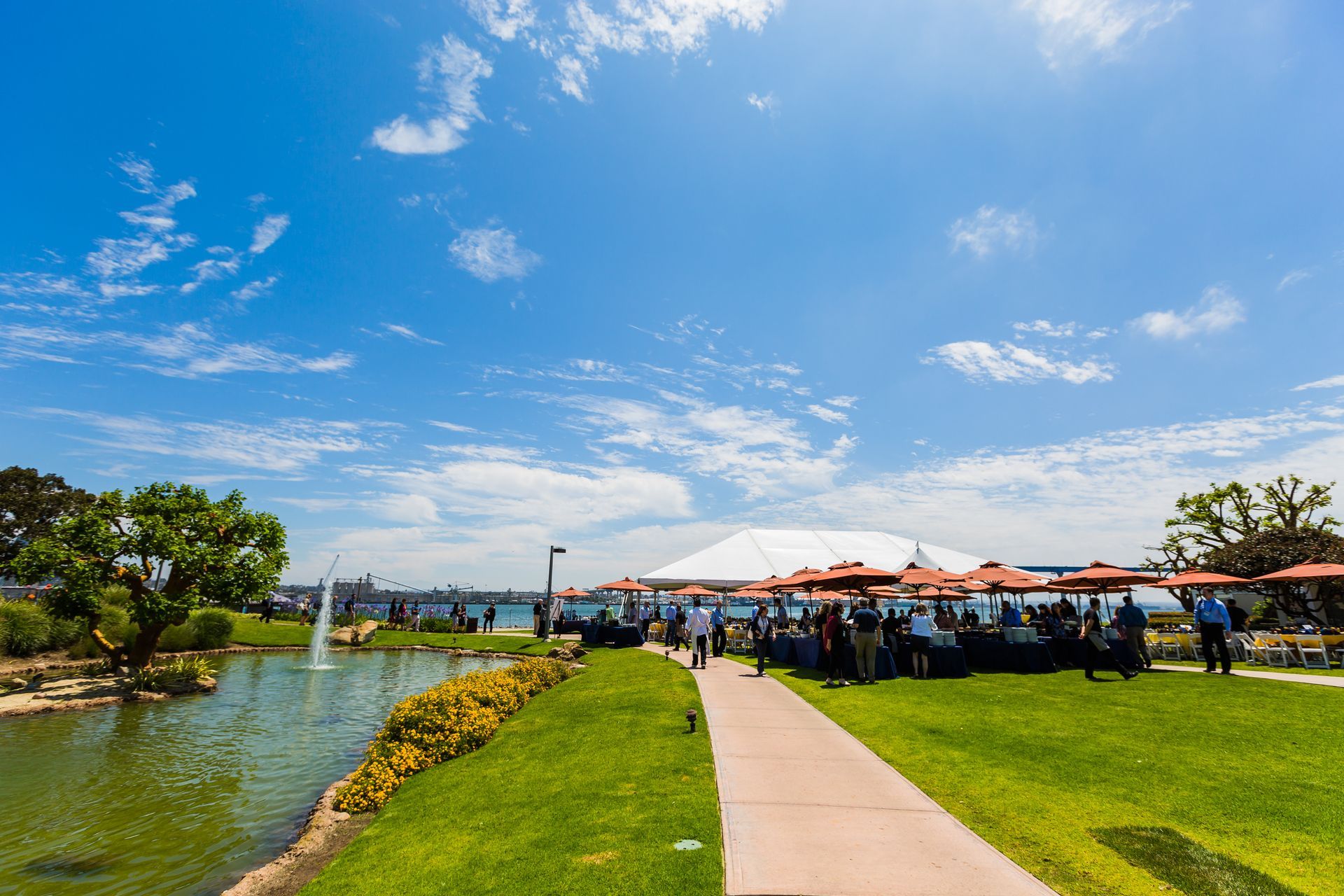 Outdoor gathering near a pond with people at tables, under umbrellas, sunny day.
