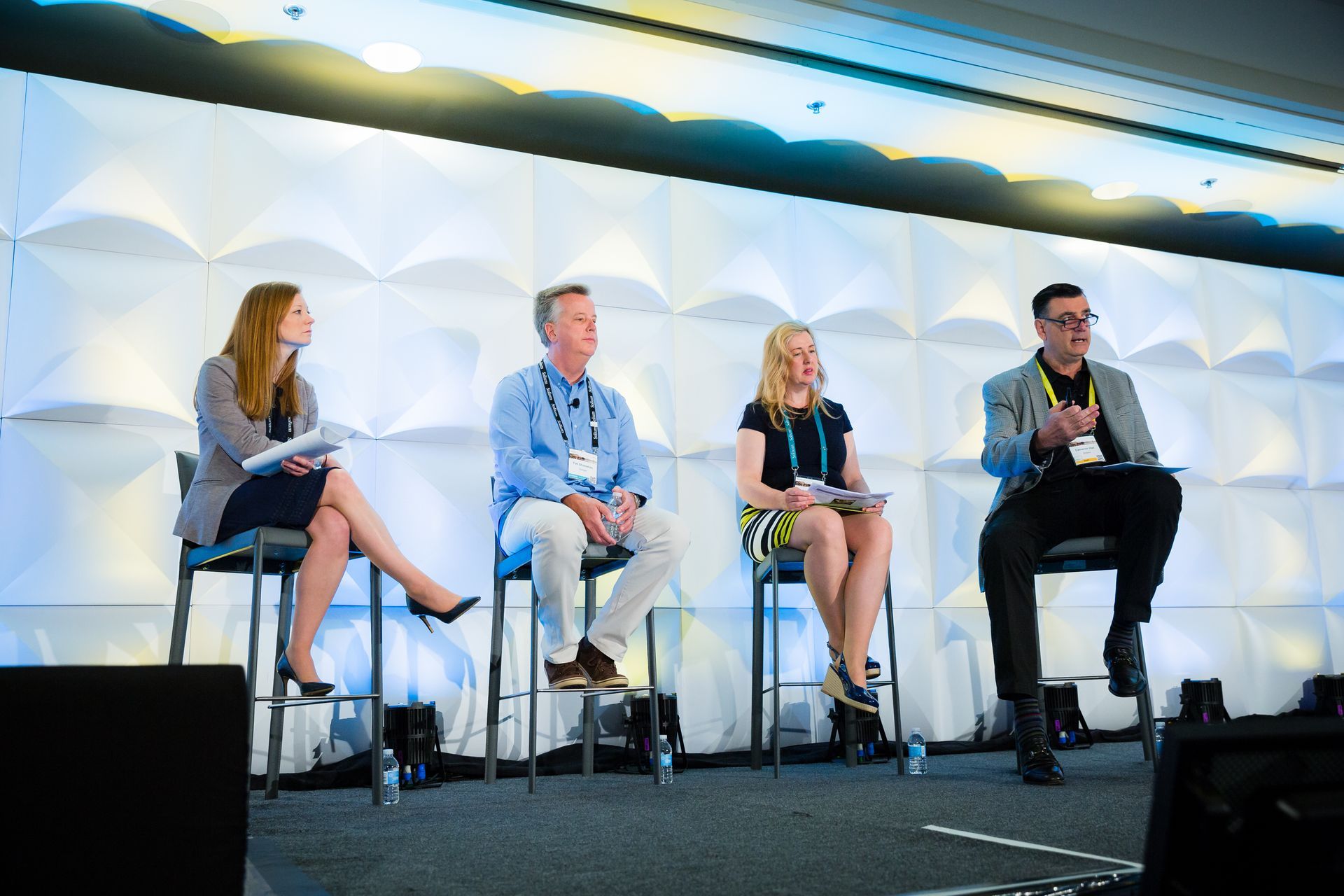 Panel of four people seated on stools, talking on a stage with a textured wall.