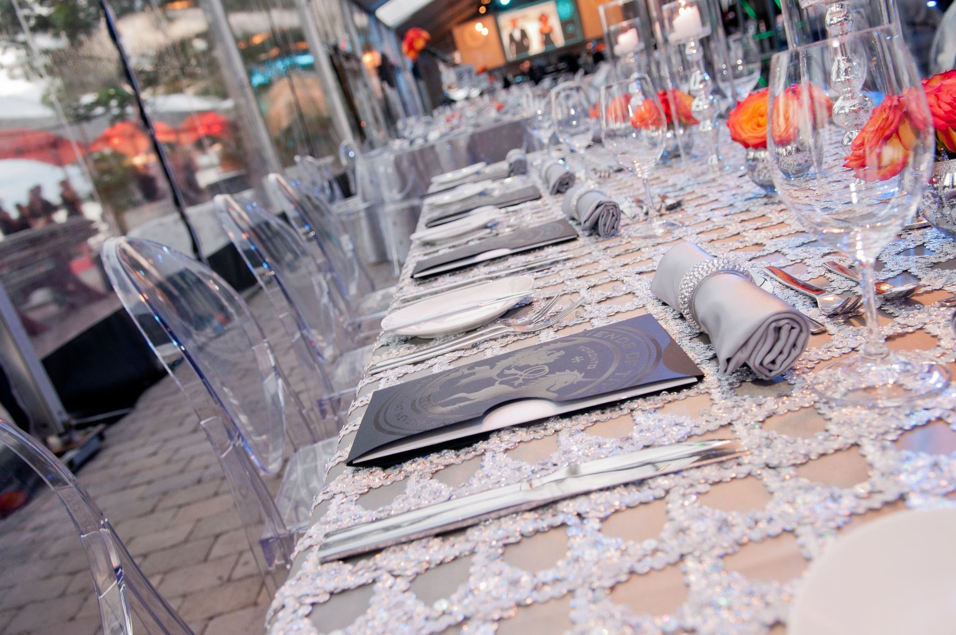 A long table with clear chairs and silverware on it.