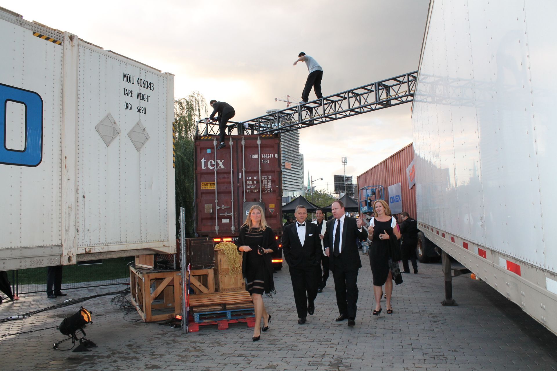 People in formal wear walking past shipping containers; men on a raised structure.