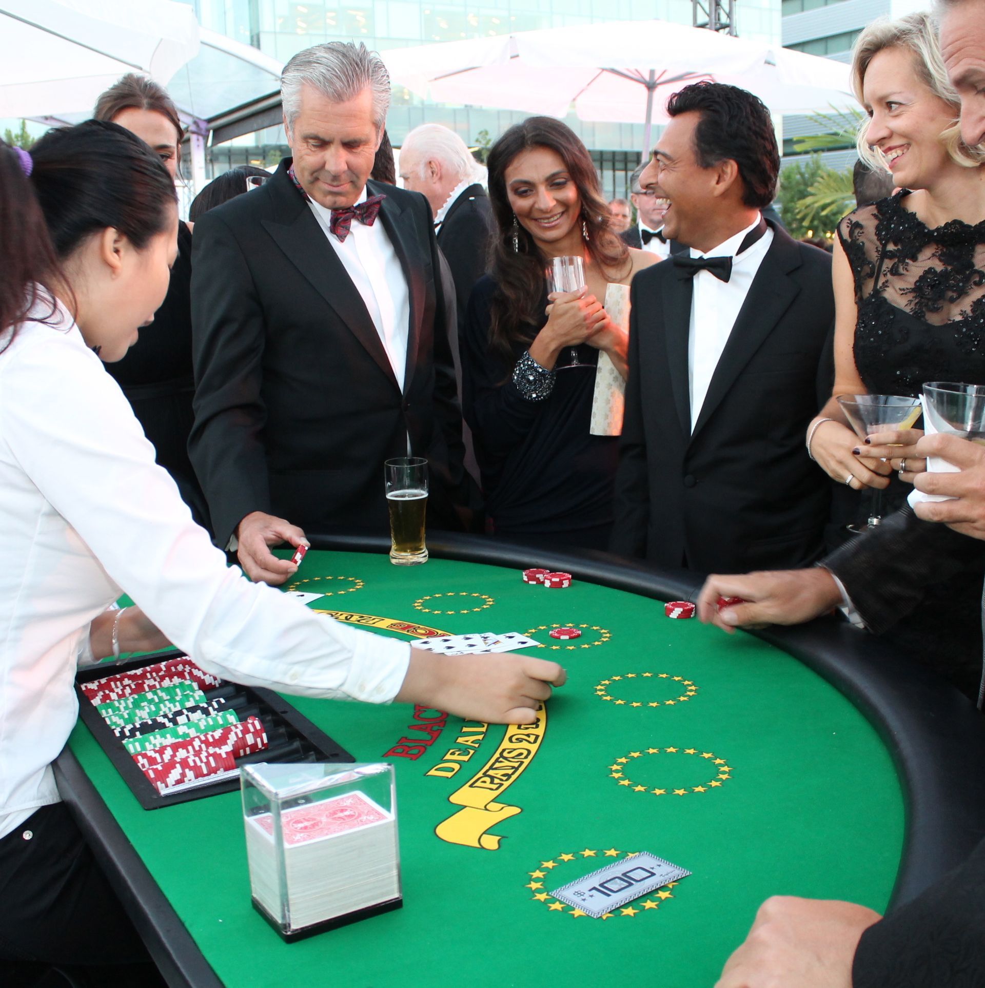 People playing blackjack at an outdoor event; dealer handling cards, others holding drinks, evening attire.