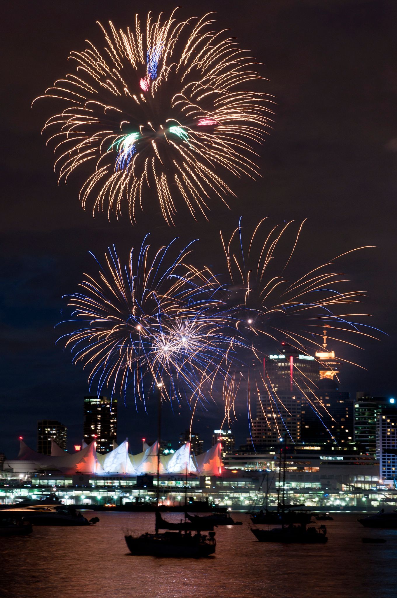 Fireworks burst over a city skyline at night, with boats in the water.