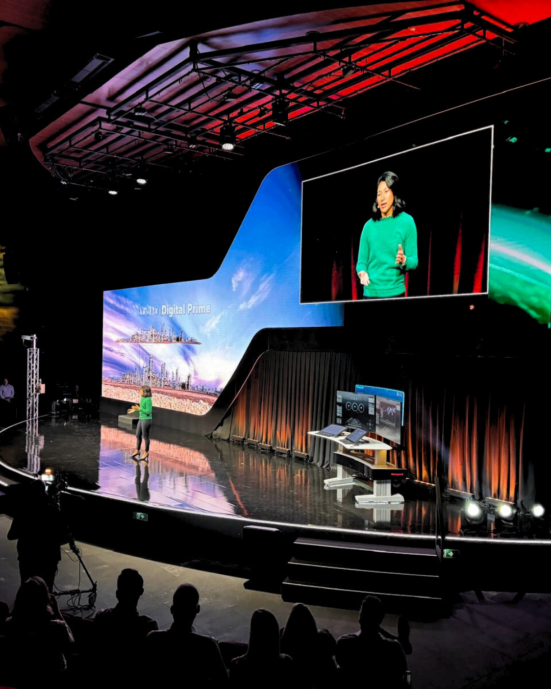 Speaker on a lit stage with large screens and audience silhouettes during a presentation