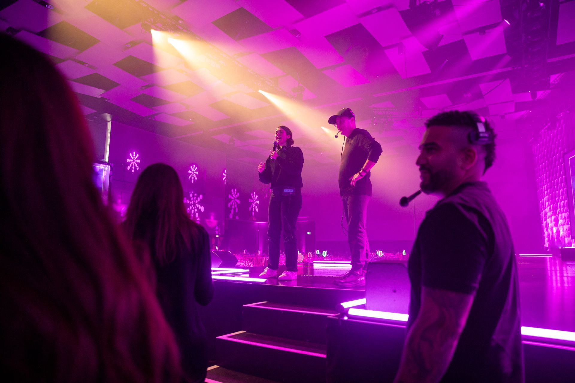 Stage with two men, one speaking into a microphone. Purple and yellow stage lighting. Audience in foreground.