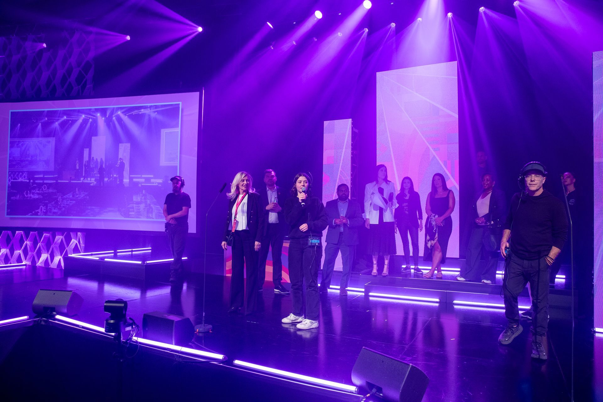 People on stage under purple stage lights, some clapping. Video screen in background.